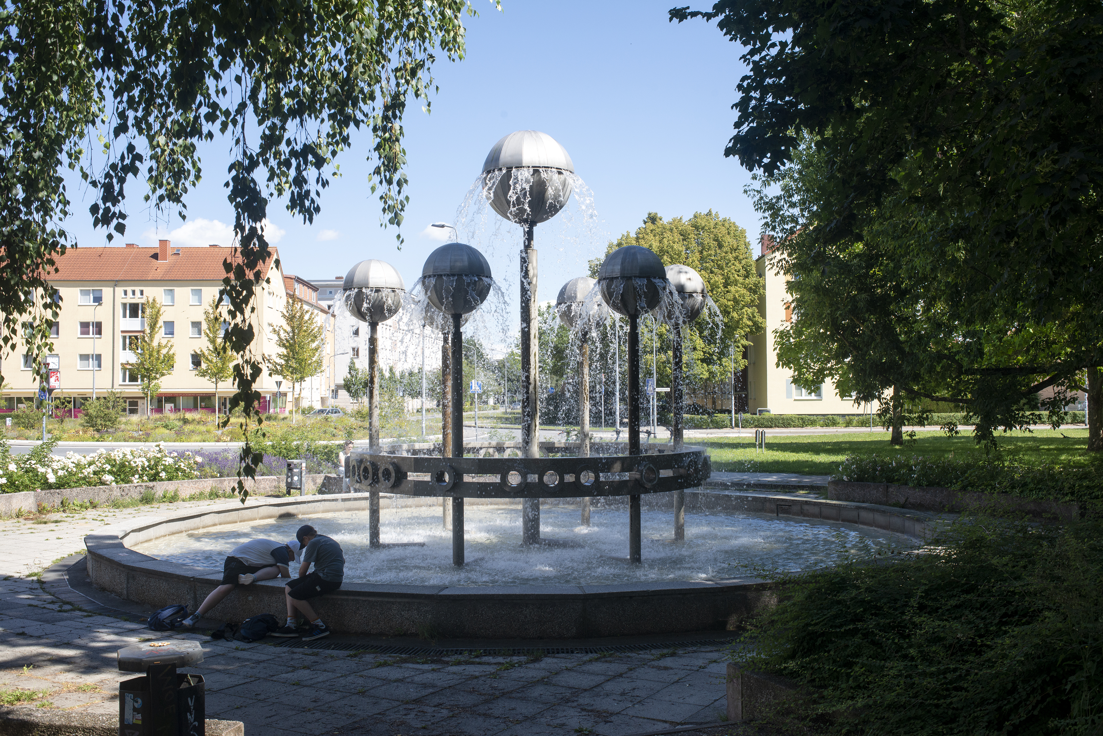 Zwischen Bäumen liegt ein kleiner Platz mit einem runden Brunnenbecken. In der Mitte steht ein Brunnen, der aus sieben Metallstäben mit Kugeln obendrauf besteht. Aus den Kugeln fließt Wasser hinunter in das Brunnenbecken.