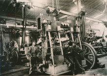 Drei Arbeiter der Neptunwerft posieren um 1925 in einer Werkshalle mit Maschinenpark.