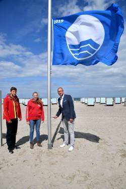 Die DRK-Rettungsschwimmer Max Cornelius und Sarah Wenzel hissen gemeinsam mit Matthias Fromm, Tourismusdirektor von Rostock & Warnemünde, die Blaue Flagge am Strand von Warnemünde 
