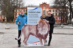 Oberbürgermeister Claus Ruhe Madsen (re.) und Zoodirektor Udo Nagel mit dem Löwen, der vor den unsichtbaren Gefahren der Corona-Pandemie warnen und für die luca-App werben soll.